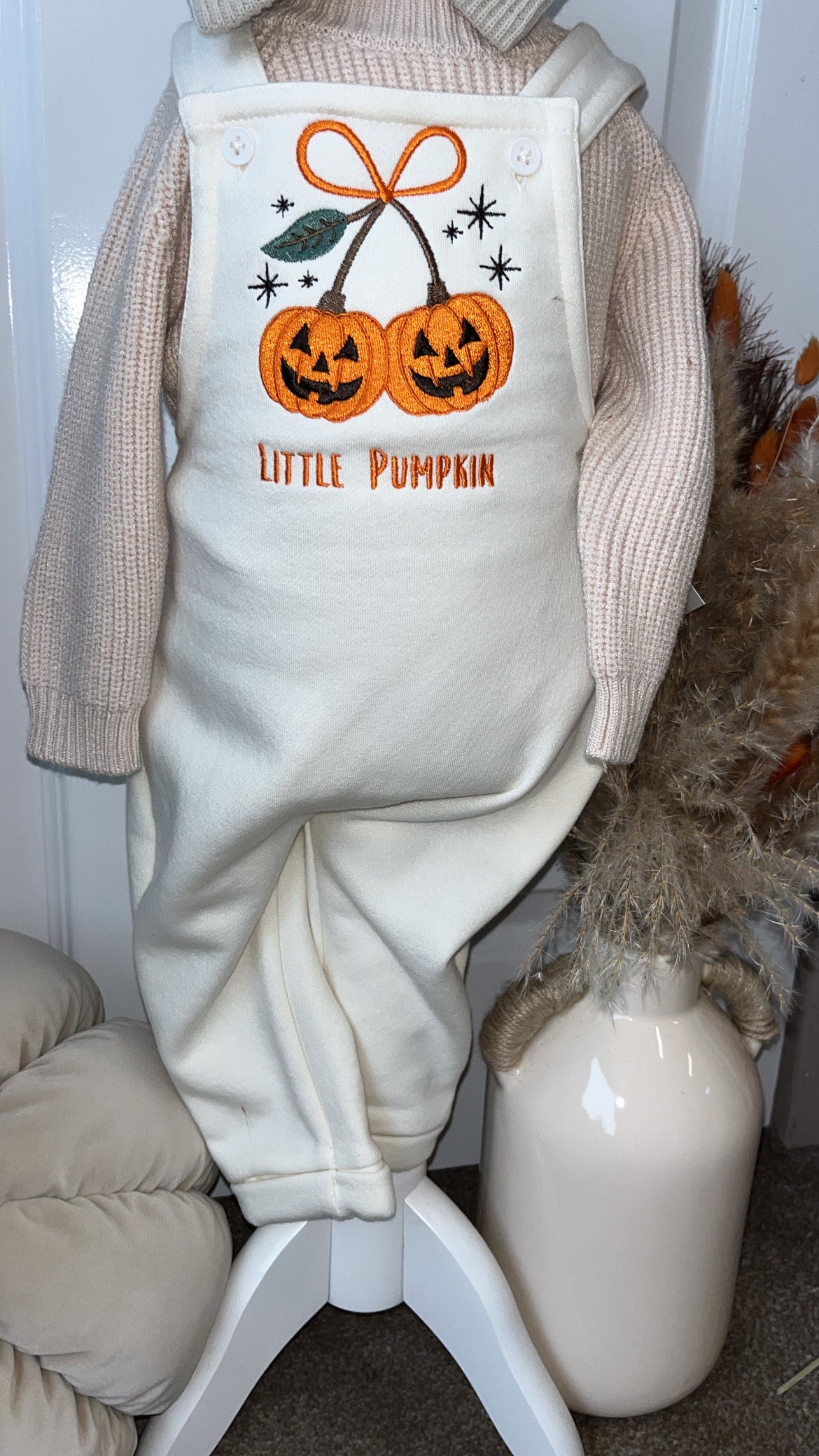 A baby or toddler knit one-piece outfit in cream with a Halloween graphic of two orange pumpkins, an orange bow, and the text 'LITTLE PUMPKIN' on the bib front, styled with a beige knit hat and standing on a white display stand.