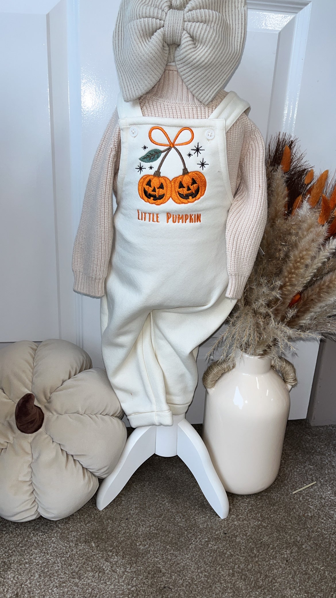 A baby outfit displayed on a mannequin or stand, featuring cream-colored overall with a bow-tie chest graphic of two jack-o'-lanterns and the words 'LITTLE PUMPKIN,' layered over a light pink knit long-sleeve top. The scene is styled with a large beige bow, a pumpkin pillow, a ceramic vase with dried grasses, and a white door in the background.