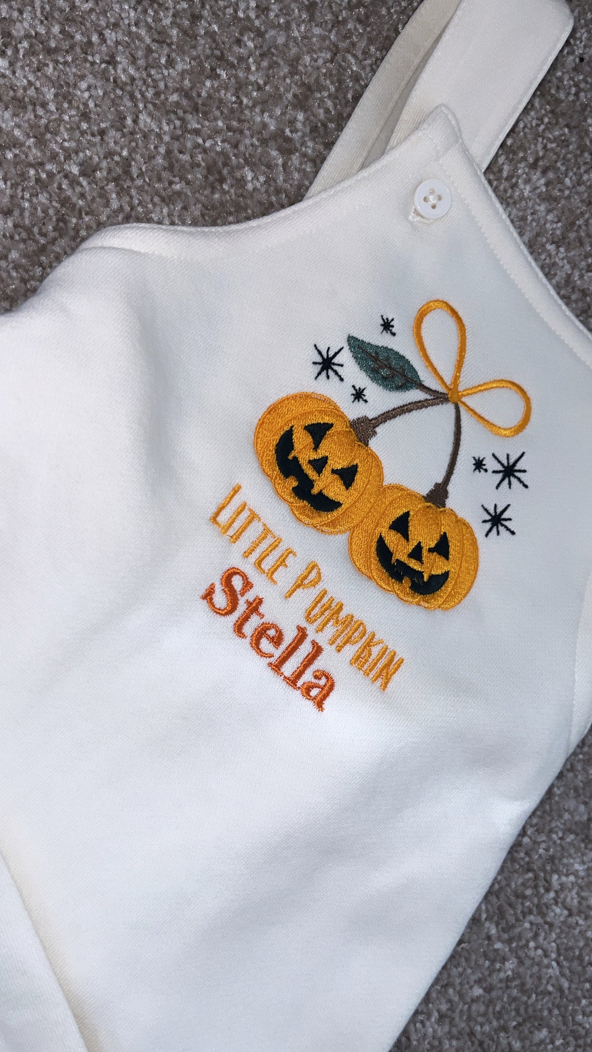 A white baby bodysuit laid flat on a gray carpet, featuring embroidered orange jack-o'-lantern pumpkins with black faces, a yellow bow, black stars, and the text 'Little Pumpkin Stella' in orange.