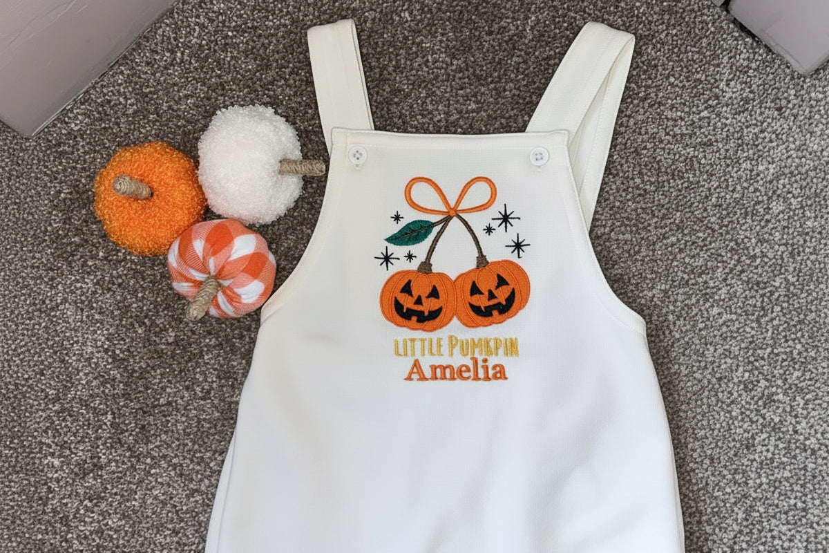 A white baby overalls romper laid flat on a gray carpet, featuring a Halloween graphic of two jack-o'-lantern pumpkins with an orange bow and the text 'LITTLE PUMPKIN Amelia' in orange. Decorative small pumpkins and a gray fabric tag are placed beside the garment.